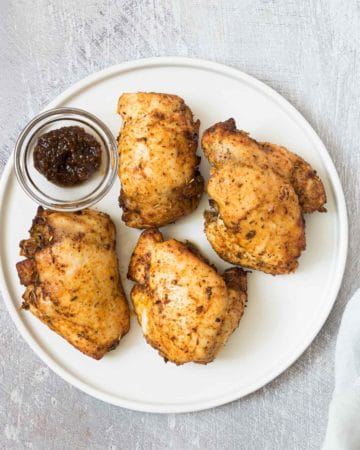 a top down view of reheat chicken thighs in air fryer that are served on a white plate with a cup of sauce
