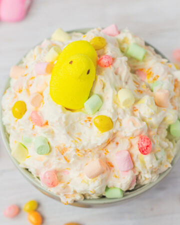 A top-down view of Easter fruit fluff in a bowl, decorated with pastel marshmallows, jelly beans, and a yellow Peeps chick, with cookies and candies scattered in the background.
