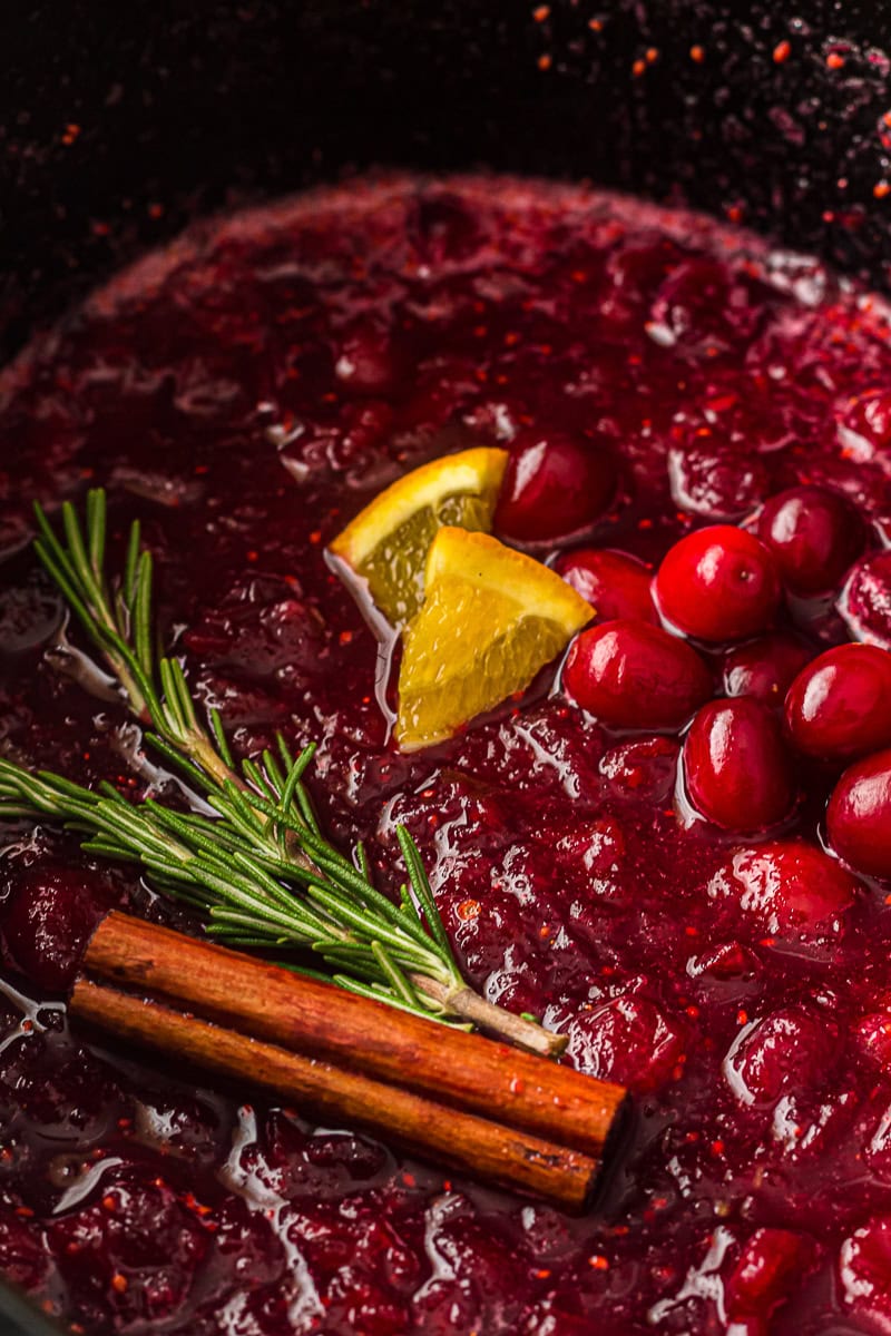 Cranberry sauce cooking in a slow cooker with orange slices, rosemary, and a cinnamon stick.