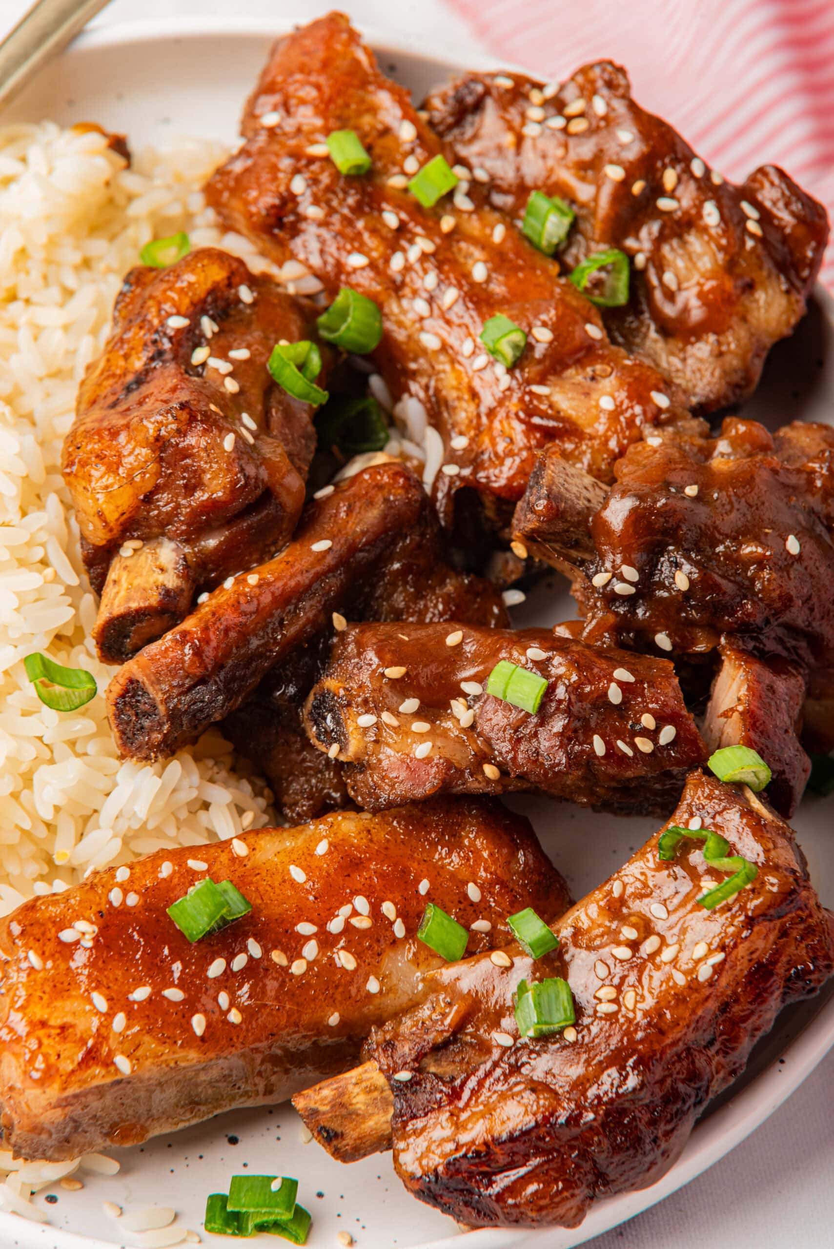 Close-up of slow-cooked ribs with a shiny glaze and sesame seeds, served over rice and garnished with green onions.
