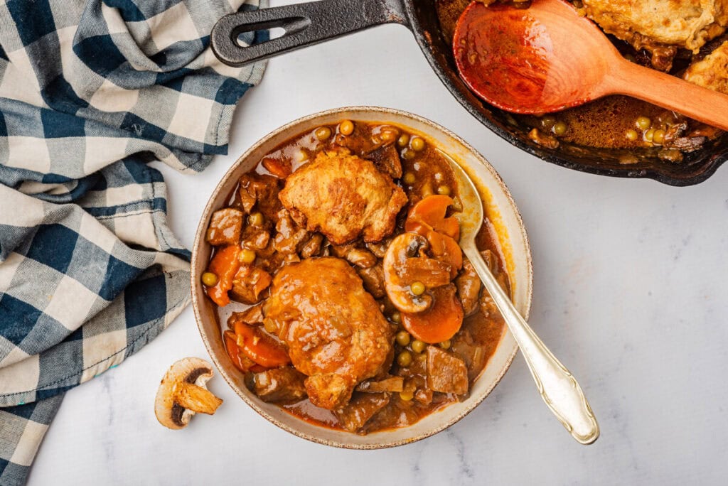 A cast-iron skillet filled with beef and vegetable stew topped with golden-brown dumplings, sitting next to a can of Guinness beer and a checkered cloth.