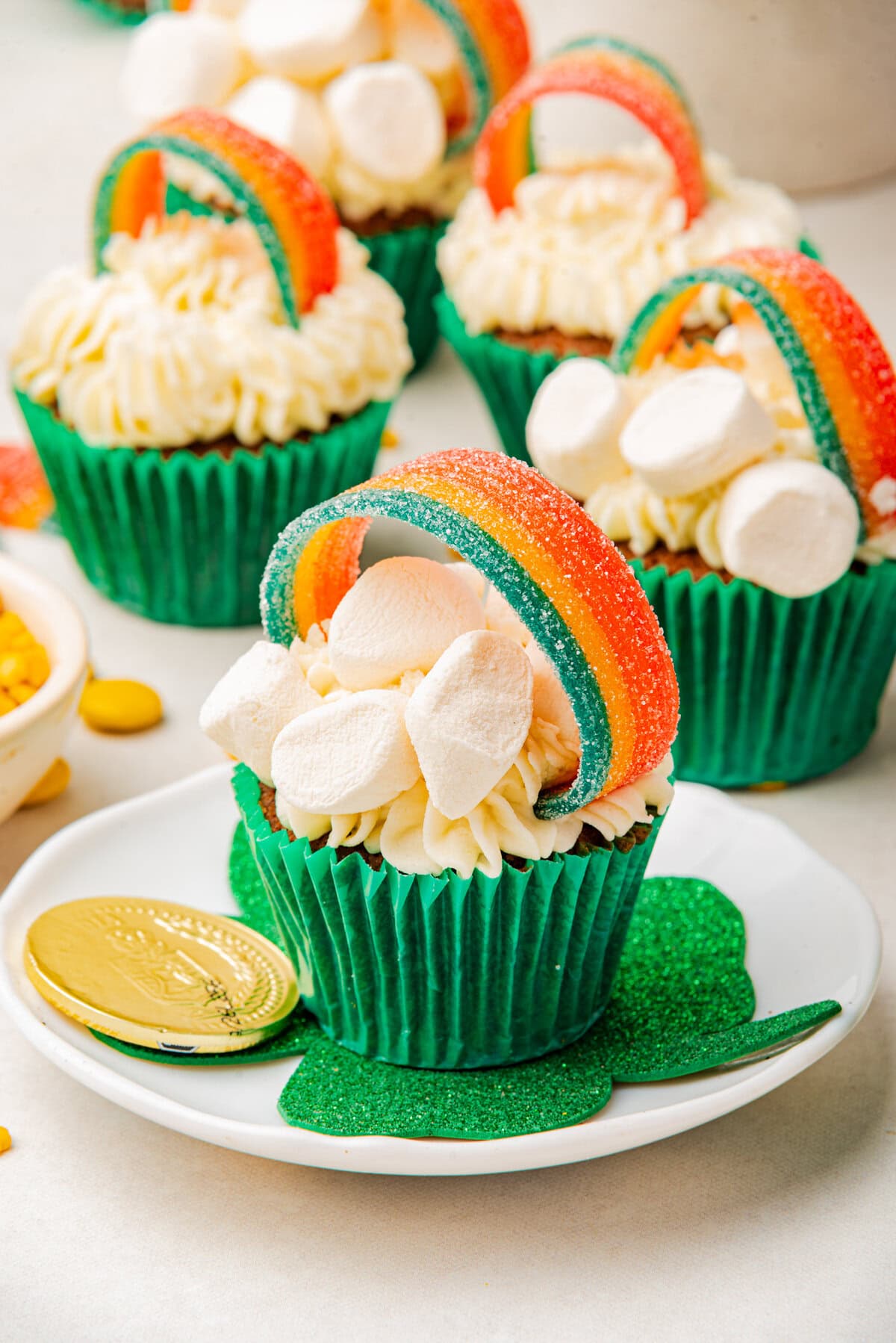 A close-up of a cupcake topped with a rainbow candy handle, marshmallows, and fluffy frosting, placed on a white plate with gold chocolate coins.