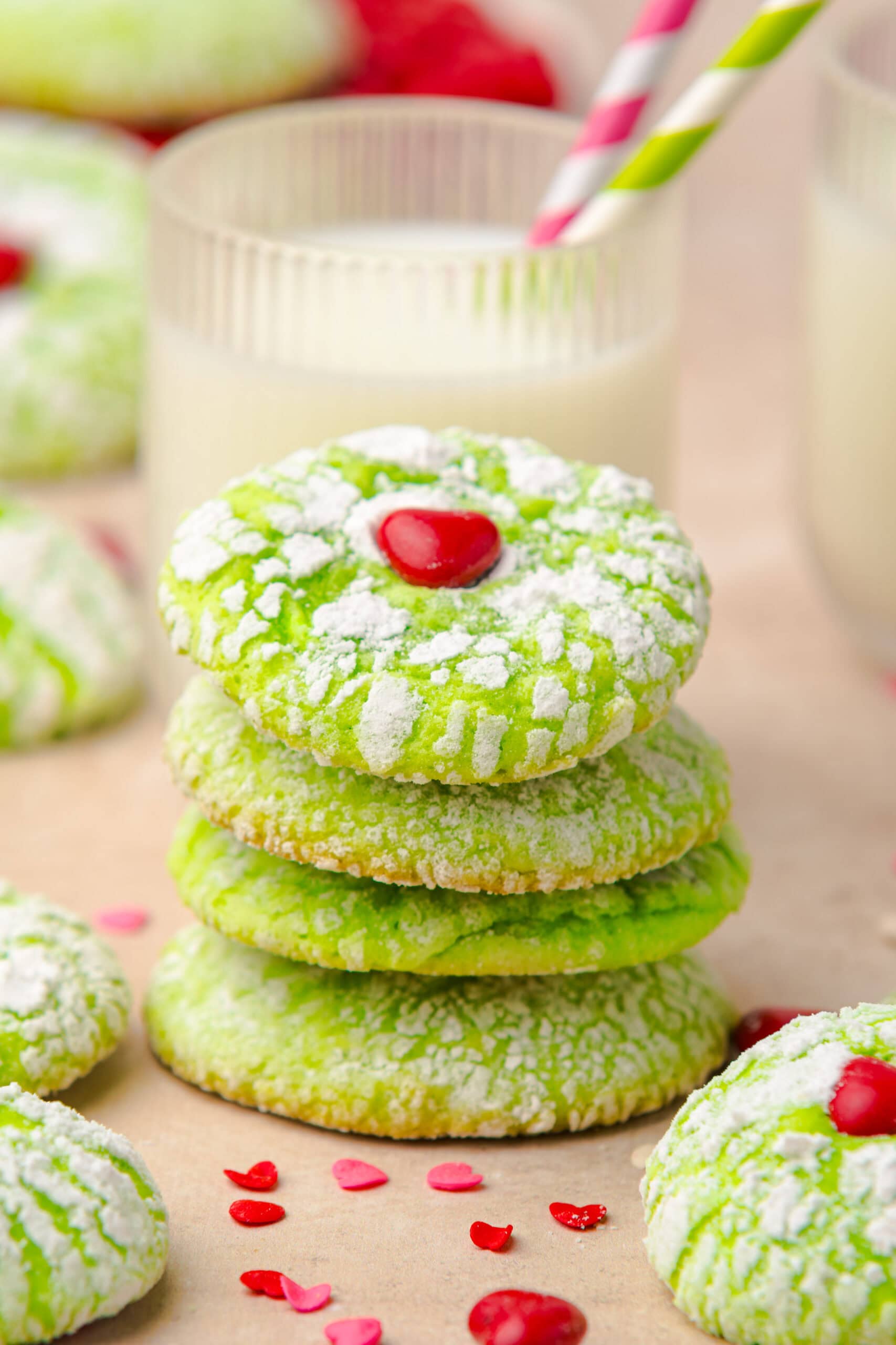 Stack of festive green cookies coated in powdered sugar, each topped with a red heart candy, with glasses of milk and striped straws in the background.