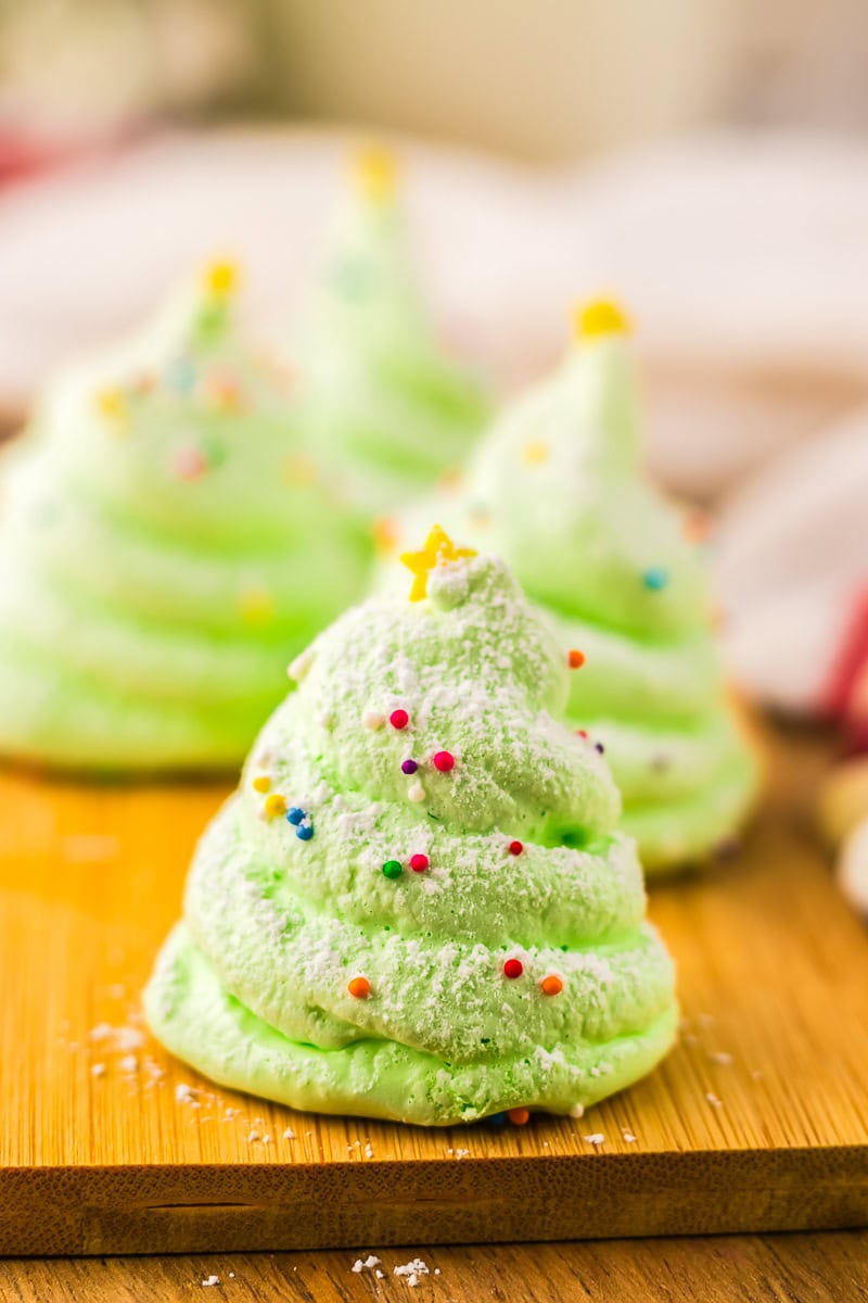 Single green Christmas tree meringue in focus, topped with a yellow sugar star and colorful sprinkles, with other decorated meringues blurred in the background.