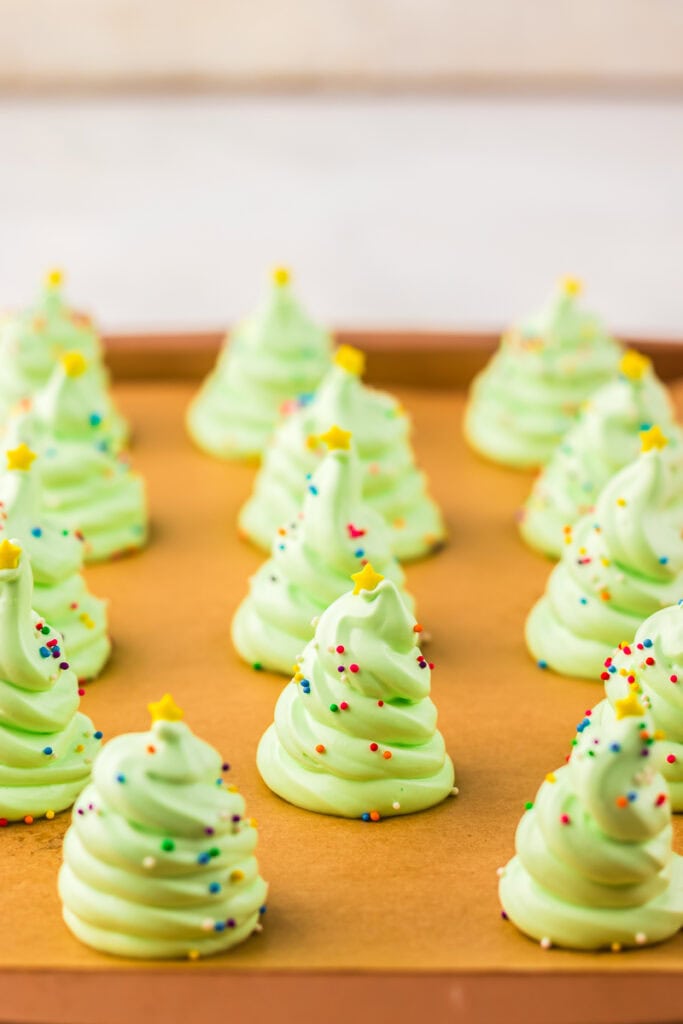 Rows of piped green meringue Christmas trees on a baking tray lined with parchment paper, each decorated with sprinkles and yellow star toppers.